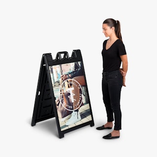 A woman wearing a black shirt, jeans and flats standing next to a black sandwich board sign.