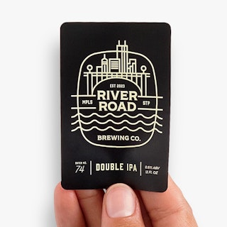 A hand holding a black and white bottle label printed with River Road Brewing Co.