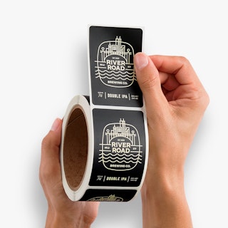 Two hands holding a roll of bottle labels printed with a black and white design and River Road Brewing Co.