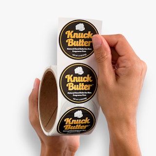 Two hands holding circle roll labels printed with a black, yellow and white design and Knuck Butter in the middle.