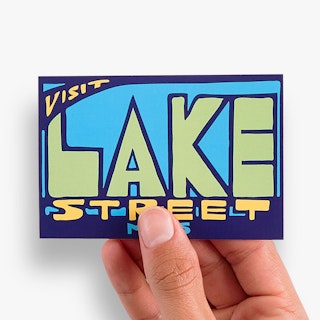 A hand holding a custom decal printed with Visit Lake Street in blue, green and yellow.