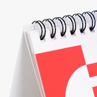 The corner of a custom desk calendar printed with a red and white design and black coil binding.