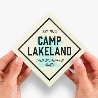 Two hands holding a die-cut magnet printed with Camp Lakeland in black with a graphic of Minnesota.