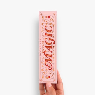 A hand holding a foil bookmark printed with Books Are Magic in pink, red and gold foil.