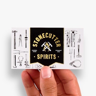A hand holding a custom business card printed with Stonecutter Spirits in gold foil on a black background.