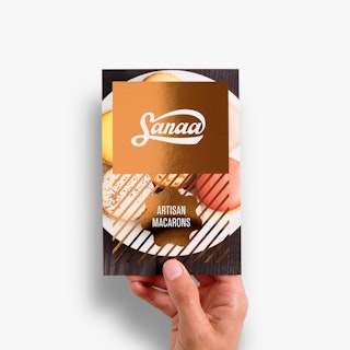 A hand holding a flat card printed with Sanaa Artisan Macarons and gold foil over an image of macarons.