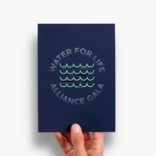 A hand holding a dark blue foil invite printed with Water For Life Alliance Gala and blue-green foil waves.