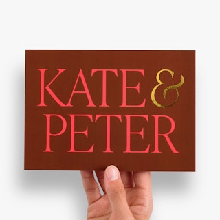 A hand holding a maroon wedding invite printed with Kate & Peter in red and gold foil.