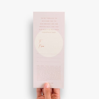 A hand holding a custom gift certificate printed with spa information and lines for To and From.