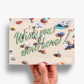 A hand holding a greeting card printed with an image of people laying on a beach and Wish You Were Here.