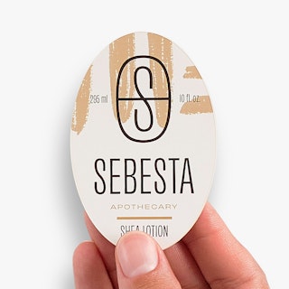 A hand holding a lotion bottle label printed with an oval shape and black and tan design.
