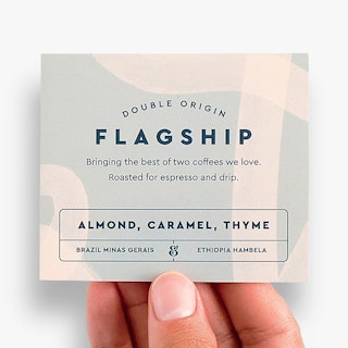 A hand holding a square product label printed with Double Origin Flagship and coffee bean details.