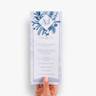 A hand holding a wedding menu card printed with a blue and white foliage design and ceremony details.