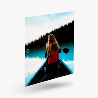 A metal print of a woman paddling in a canoe on a lake with a forest of trees in front of her.