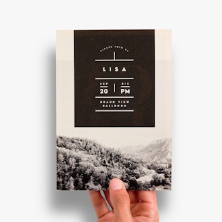 A hand holding a printed invitation with a black and white forest design and gala details on the back.