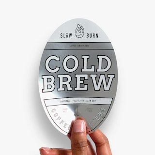A hand holding an oval-shaped label printed with Slow Burn Cold Brew in silver, black and white.