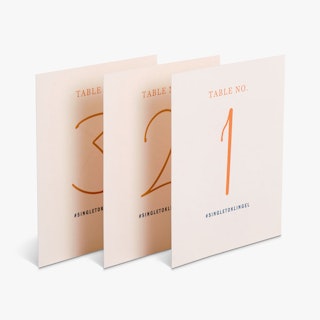 Three custom place cards printed with a light pink background, peach table numbers and a black hashtag.