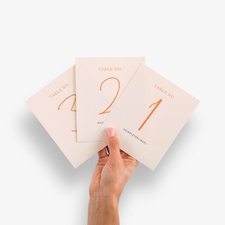 A hand holding three place cards fanned out and printed with a pink background and orange table numbers.