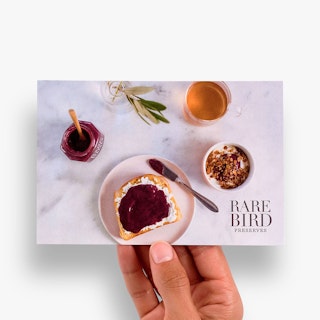 A hand holding a marketing postcard printed with Rare Bird Preserves and a piece of toast on a white plate.