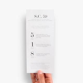 A hand holding a custom rack card printed with S.C. 59 and perfume scent details.