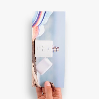 A hand holding a business rack card printed with a perfume bottle in blue, white, brown and orange.