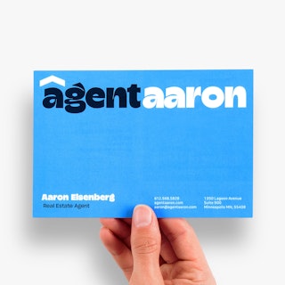 A hand holding a blue realtor postcard printed with Agent Aaron and contact information.