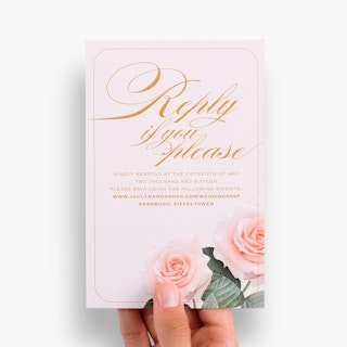 A hand holding a wedding reception card printed with Reply If You Please in cursive orange text.