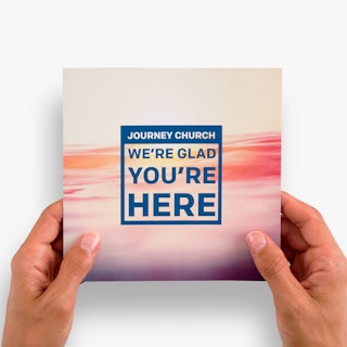 Two hands holding a church welcome brochure printed with We're Glad You're Here in blue on the cover.