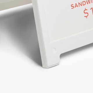 The bottom corner of a white sidewalk sign holding a printed sign in white and red.