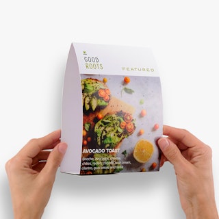 Two hands holding a restaurant table tent printed with avocado toast and Good Roots in black and green.