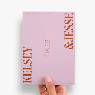 A hand holding a save the date card with a pink background and Kelsey & Jesse in dark orange.