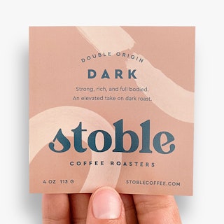 A hand holding a square product label printed with Double Origin Dark and Stoble Coffee Roasters.