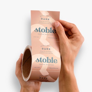 Two hands holding a roll of square labels printed with Double Origin Dark and details about coffee beans.