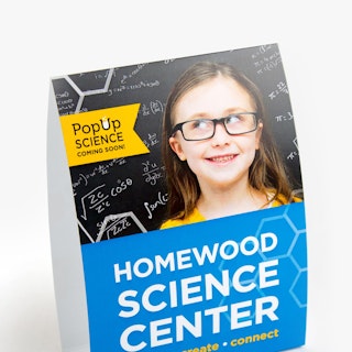 A table tent advertising a science event with a girl in glasses in front of a chalkboard.
