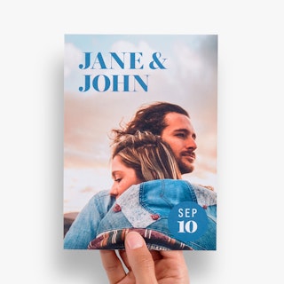 A hand holding a custom wedding invitation printed with Jane & John and a man and woman hugging.