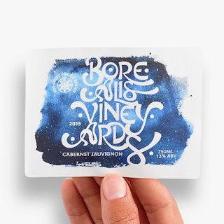 A hand holding a custom wine label printed with Borealis Vineyards in blue, white and black.